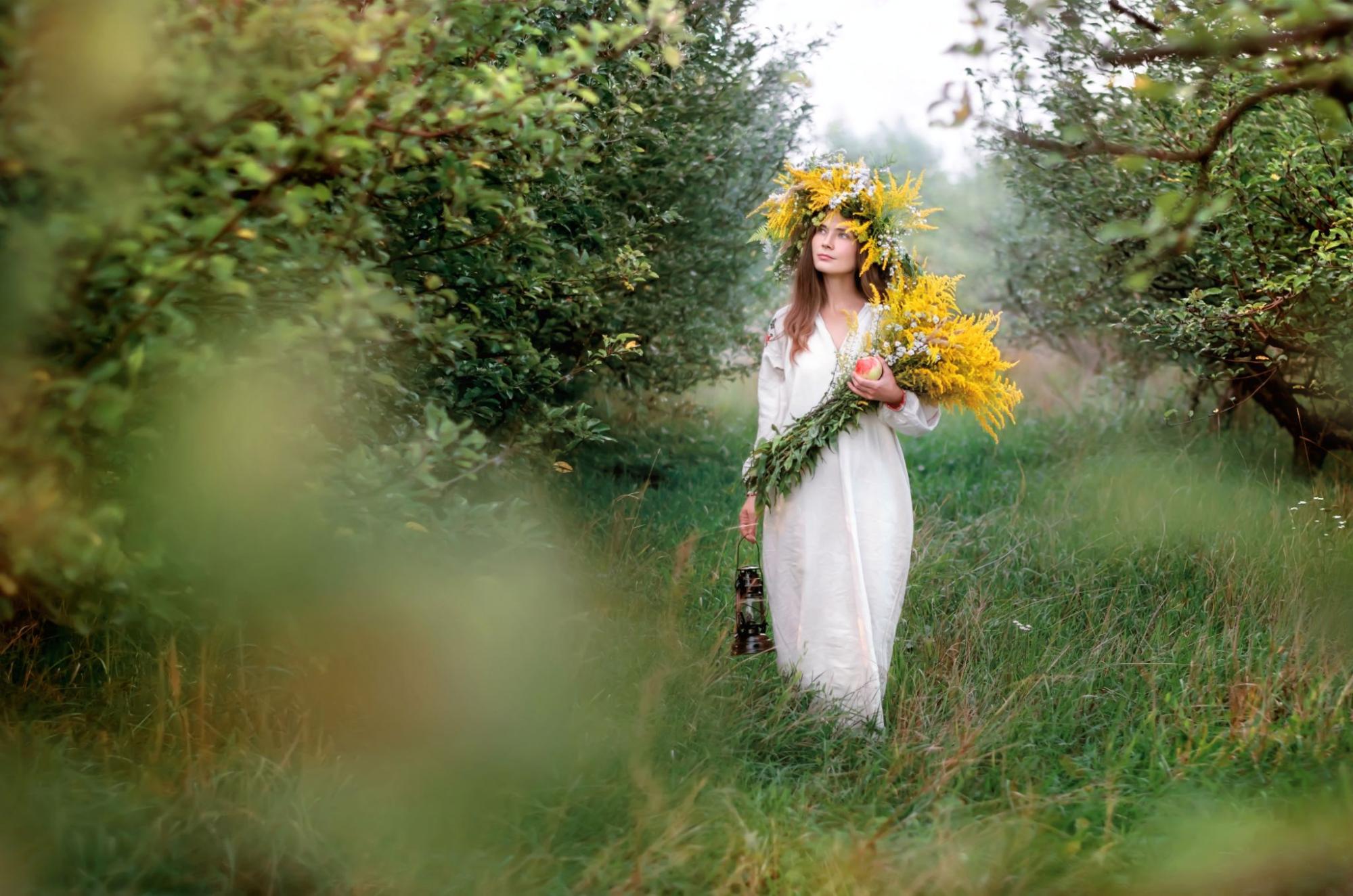 A young woman in a wreath and a long white national shirt collects wildflowers